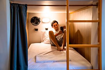 Viajero Hostels | Viajero Medellín | Chico cargando su celular en el tomacorriente de su cubiculo personal, con luz de lectura y ventilador en el hostal en el poblado Medellin