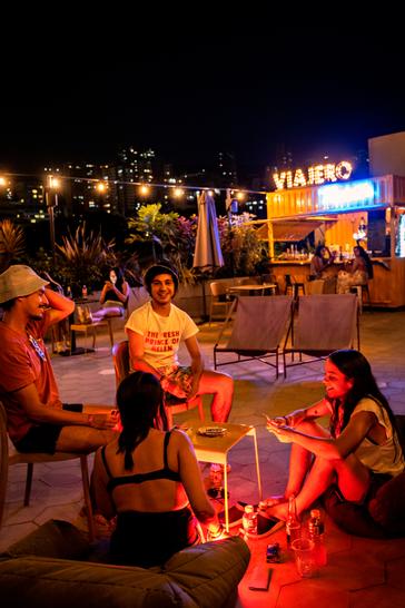 Viajero Hostels | Viajero Medellín | Grupo de amigos compartiendo mientras juegan cartas en el rooftop de Viajero hostel Medellin con luces, comódos sofás y bar